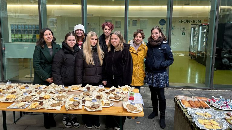 Eine Gruppe von Frauen steht hinter einem Tisch mit einer Vielzahl veganer Desserts, einschließlich Kekse und Kuchen, alle als vegan gekennzeichnet. Sie lächeln und posieren für ein Foto.