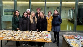 Eine Gruppe von Frauen steht hinter einem Tisch mit einer Vielzahl veganer Desserts, einschließlich Kekse und Kuchen, alle als vegan gekennzeichnet. Sie lächeln und posieren für ein Foto.