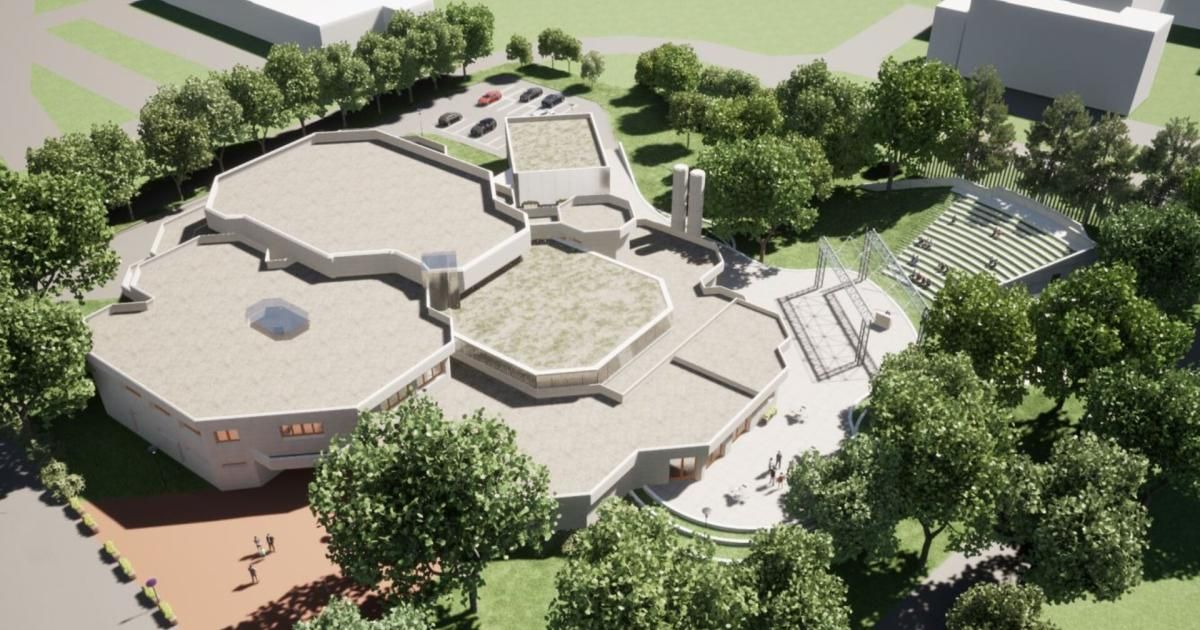 Ein Blick von oben auf ein modernes Gebäude mit sechseckigen Formen, umgeben von grünem Gras und Bäumen. Mehrere Autos sind auf dem Parkplatz geparkt. Menschen gehen herum.