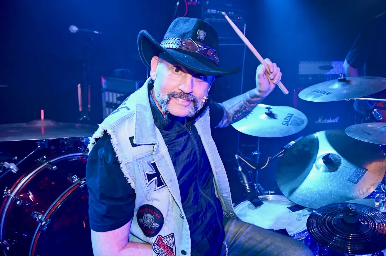 Ein Mann mit einem Schnurrbart und Tattoos am Arm spielt Schlagzeug auf der Bühne. Er trägt einen schwarzen Hut und eine Weste mit Aufnähern.