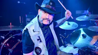 Ein Mann mit einem Schnurrbart und Tattoos am Arm spielt Schlagzeug auf der Bühne. Er trägt einen schwarzen Hut und eine Weste mit Aufnähern.