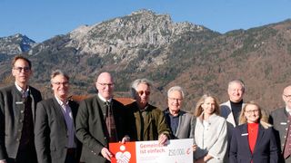 Gruppe von Personen, die einen großen Scheck vor Bergen halten, wobei eine Person ein Schild mit der Aufschrift Gemeinsam ist einfach. 250.000 Euro. in der Hand hält.