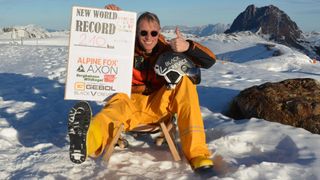 Ein Mann sitzt auf einem Schlitten in verschneiten Bergen und hält ein Schild für einen neuen Weltrekord. Er trägt ein gelbes Outfit, eine Schutzbrille und einen Helm. Hinter ihm sind Berge schneebedeckt.
