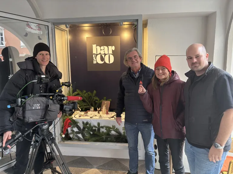 Vier Personen stehen in einem Raum mit einer Kamera und einem Stativ und lächeln. Das Schild darüber lautet 'bar & co'. Hinter ihnen ist ein Tisch mit Weihnachtsdekorationen geschmückt.