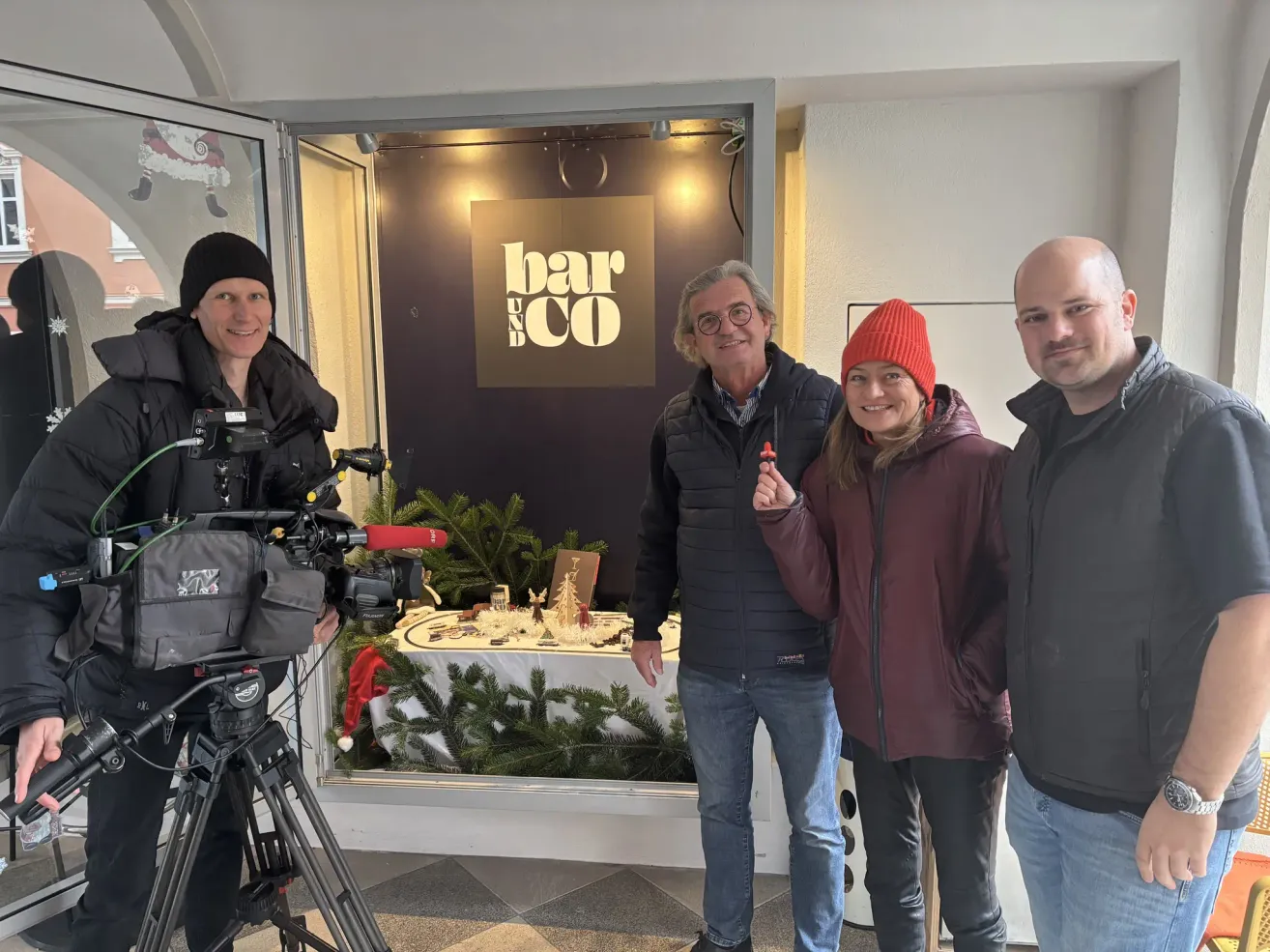 Vier Personen stehen in einem Raum mit einer Kamera und einem Stativ und lächeln. Das Schild darüber lautet 'bar & co'. Hinter ihnen ist ein Tisch mit Weihnachtsdekorationen geschmückt.
