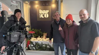Vier Personen stehen in einem Raum mit einer Kamera und einem Stativ und lächeln. Das Schild darüber lautet 'bar & co'. Hinter ihnen ist ein Tisch mit Weihnachtsdekorationen geschmückt.
