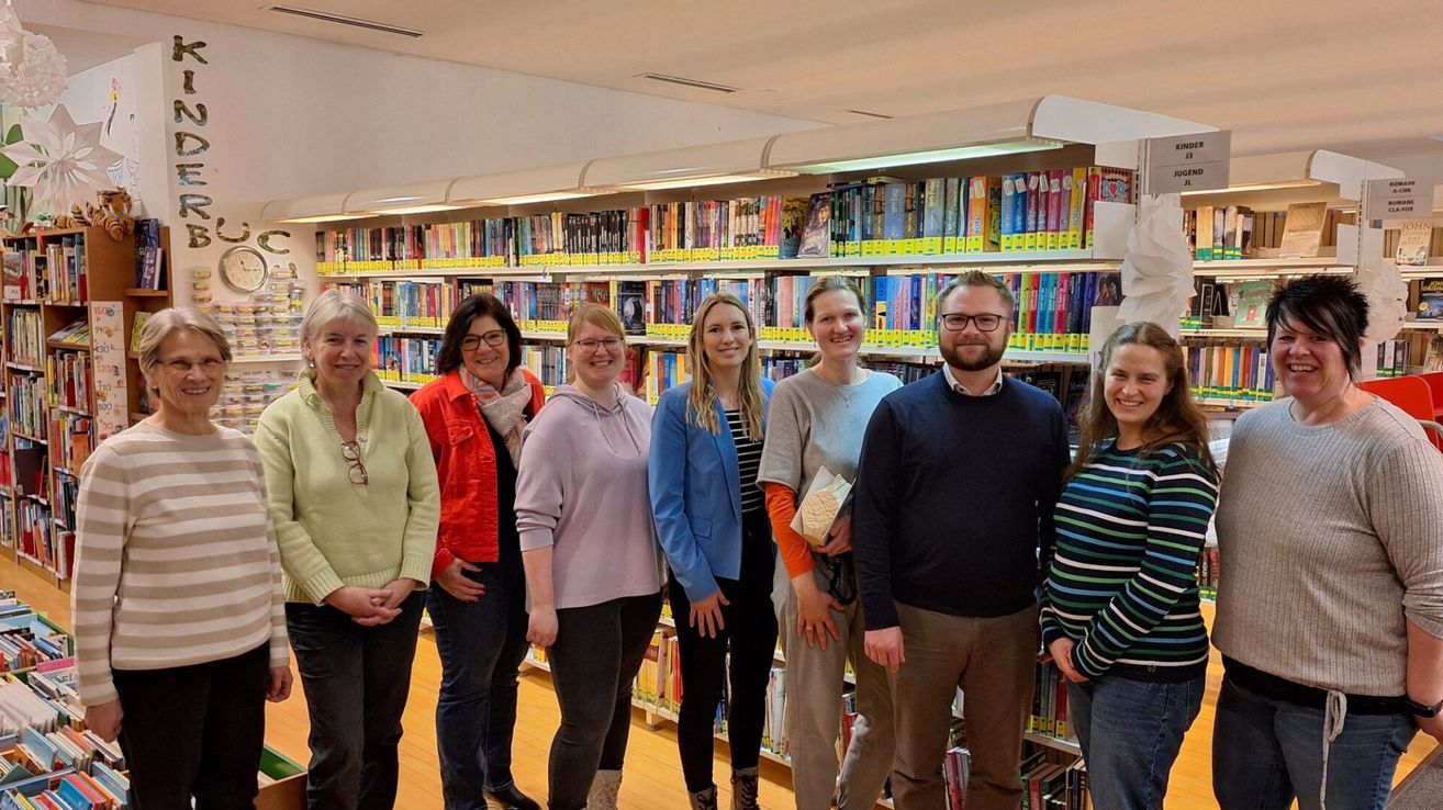 Eine Gruppe von Erwachsenen steht in einer Bibliothek, lächelt und posiert für ein Foto. Hinter ihnen sind Regale mit bunten Büchern gefüllt.