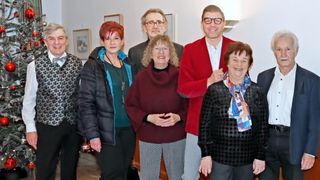 Eine Gruppe von Erwachsenen, drei Frauen und zwei Männer, stehen eng beieinander. Sie lächeln für ein Foto. Die Frau auf der linken Seite trägt eine schwarze Jacke. Die Frau in der Mitte trägt ein kastanienbraunes Rollkragenshirt. Der Mann auf der rechten Seite trägt eine rote Jacke.
