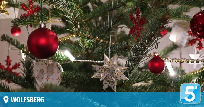 Ein geschmückter Weihnachtsbaum mit roten Kugeln, goldenem Tinsel und einem sternförmigen Ornament. Der Baum steht vor einem weißen Hintergrund mit einem blauen Banner, das '5' und ein Logo für 'WOLFSBURG' zeigt.