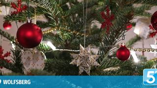 Ein geschmückter Weihnachtsbaum mit roten Kugeln, goldenem Tinsel und einem sternförmigen Ornament. Der Baum steht vor einem weißen Hintergrund mit einem blauen Banner, das '5' und ein Logo für 'WOLFSBURG' zeigt.