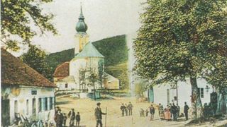 Ein altes Foto zeigt ein kleines Dorf mit einer Kirche, die einen grünen Turmhelm hat. Menschen sind auf dem Dorfplatz versammelt, mit Bäumen und Gebäuden im Hintergrund.