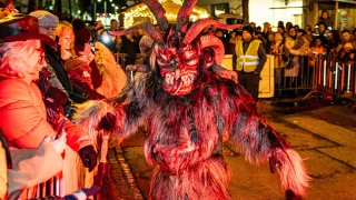 Eine Person in einem roten Teufelskostüm mit Hörnern steht vor einer Menschenmenge und hält eine Besen. Die Menge, beleuchtet von Lichtern, beobachtet von hinter einer Barriere.