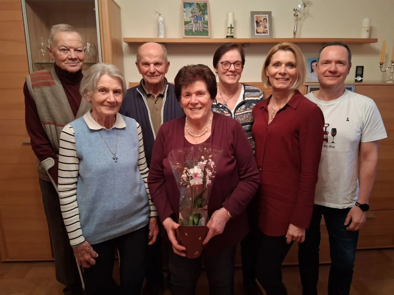 Eine Gruppe älterer Menschen steht lächelnd zusammen. Eine Frau in der Mitte hält einen Blumenstrauß. Hinter ihnen ist ein Regal mit Bilderrahmen und einer Kerze zu sehen.