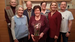 Eine Gruppe älterer Menschen steht lächelnd zusammen. Eine Frau in der Mitte hält einen Blumenstrauß. Hinter ihnen ist ein Regal mit Bilderrahmen und einer Kerze zu sehen.