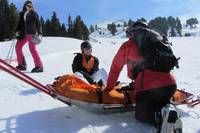 Drei Personen befinden sich in einem verschneiten Gebiet, eine Person sitzt auf einer Trage, während die anderen stehen. Sie sind alle in Winterkleidung gekleidet. Die sitzende Person wird von einer anderen Person unterstützt.