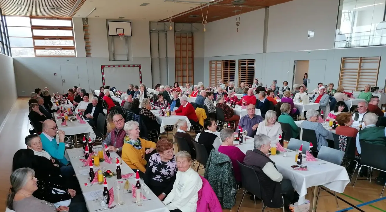 Ein großer Raum mit mehreren Tischen ist mit vielen Menschen gefüllt, die sitzen und plaudern. Die Tische sind mit Weihnachtsdekorationen geschmückt, einschließlich Flaschen mit Weihnachtsmannmützen. Der Raum hat einen Basketballkorb, eine Holzwand und Lampen.