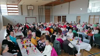 Ein großer Raum mit mehreren Tischen ist mit vielen Menschen gefüllt, die sitzen und plaudern. Die Tische sind mit Weihnachtsdekorationen geschmückt, einschließlich Flaschen mit Weihnachtsmannmützen. Der Raum hat einen Basketballkorb, eine Holzwand und Lampen.