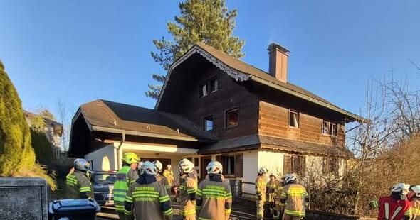 Feuerwehrleute in Schutzkleidung stehen vor einem Holzhaus mit Schornstein. Sie bereiten sich darauf vor, das Gebäude zu betreten.