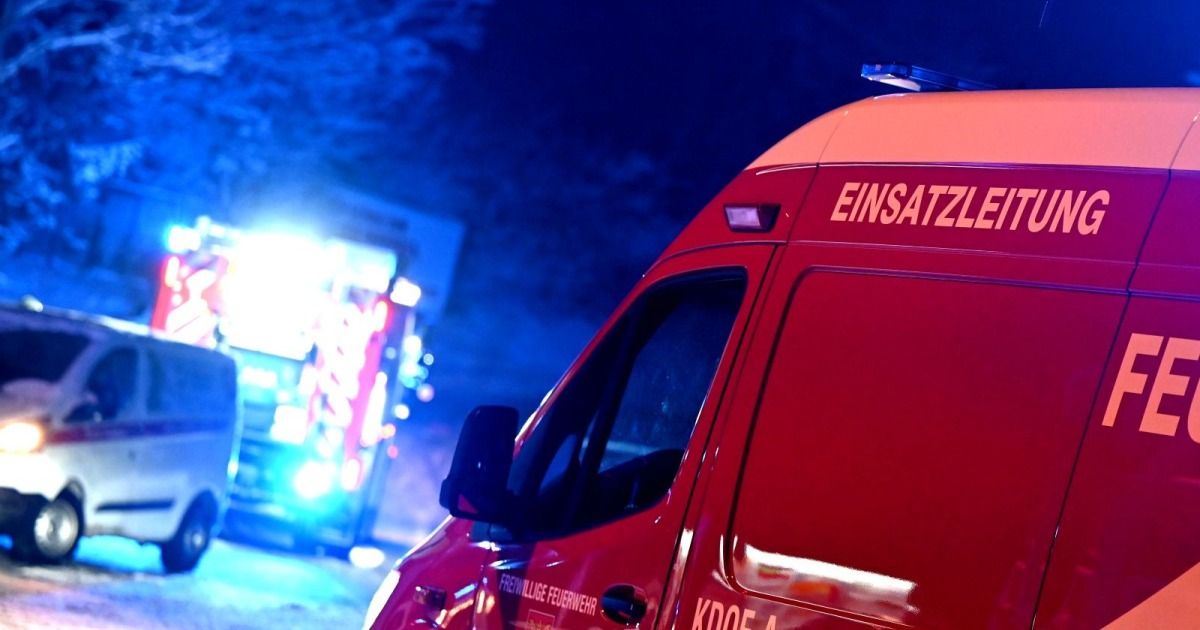 Ein rotes Einsatzfahrzeug, beschriftet mit 'EINSATZ,' steht unter einer Straßenlaterne. Die Rückseite hat sichtbare Text und Zahlen. Ein Schild mit blauen Lichtern ist im Hintergrund.