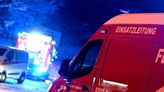 Ein rotes Einsatzfahrzeug, beschriftet mit 'EINSATZ,' steht unter einer Straßenlaterne. Die Rückseite hat sichtbare Text und Zahlen. Ein Schild mit blauen Lichtern ist im Hintergrund.