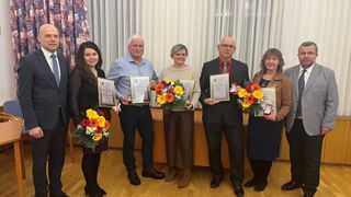 Fünf Personen stehen zusammen, halten Urkunden und Blumen, möglicherweise bei einer Preisverleihung. Eine Frau hält einen Blumenstrauß, und ein Mann auf der rechten Seite hält eine gerahmte Urkunde.