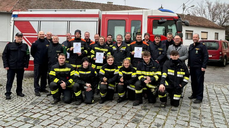 Eine Gruppe von Feuerwehrleuten in Uniformen posiert vor einem Feuerwehrauto und hält Urkunden in der Hand.