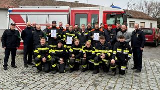 Eine Gruppe von Feuerwehrleuten in Uniformen posiert vor einem Feuerwehrauto und hält Urkunden in der Hand.