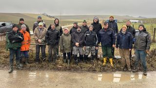 Eine Gruppe von Personen in Winterkleidung posiert für ein Foto auf einem schlammigen Feld, mit Hügeln und einer unbefestigten Straße im Hintergrund.