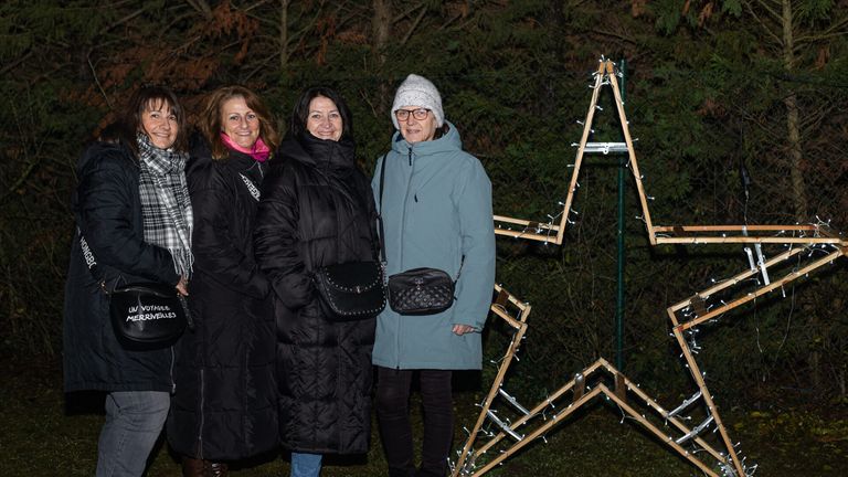 Drei Frauen stehen in Wintermänteln zusammen, lächeln und posieren für ein Foto vor einer weihnachtsbaumförmigen Dekoration.