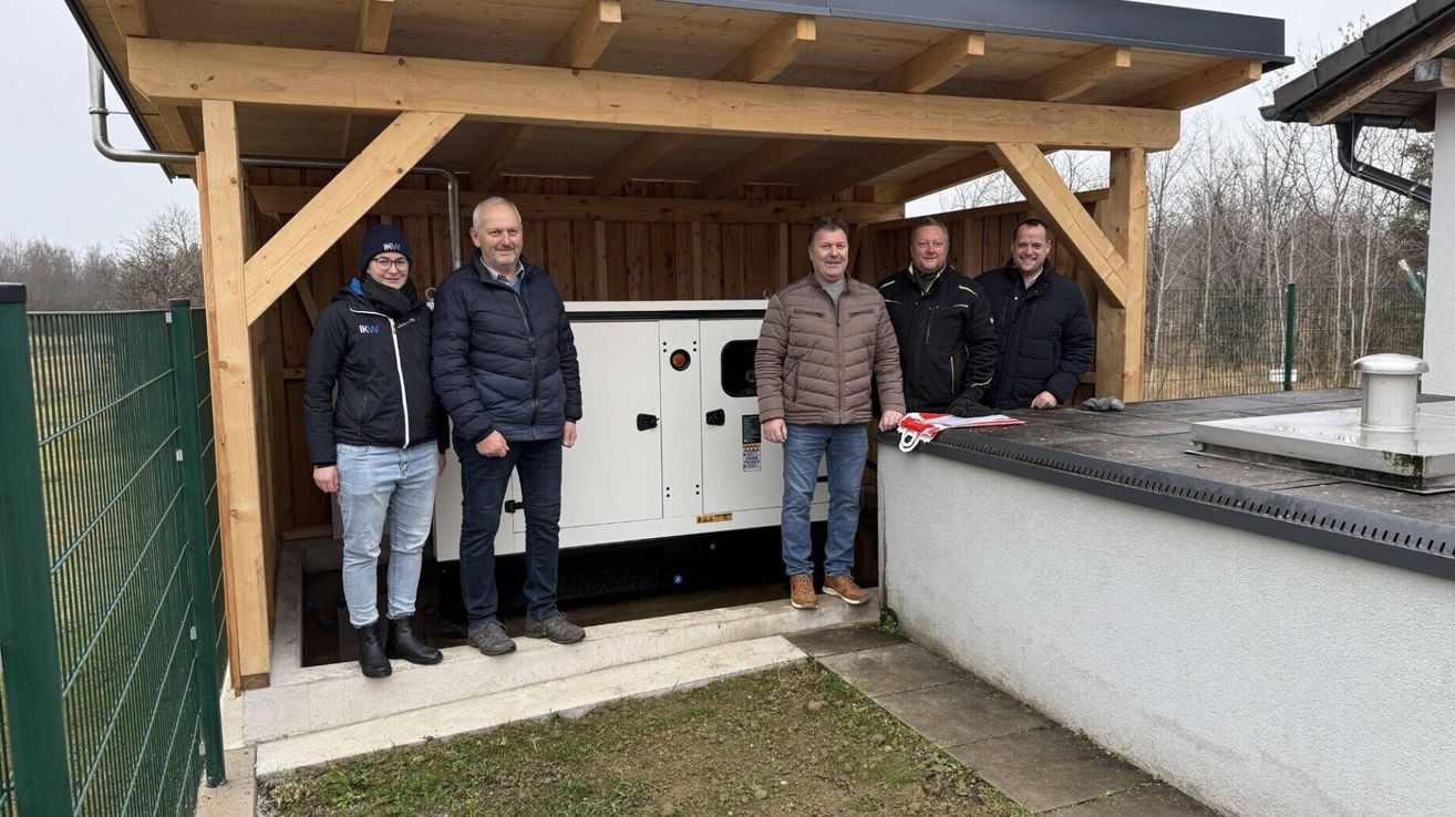 Fünf Männer stehen vor einem Gebäude mit einem Holzdach und zweien mit Brille. Sie lächeln und scheinen für ein Foto zu posieren.