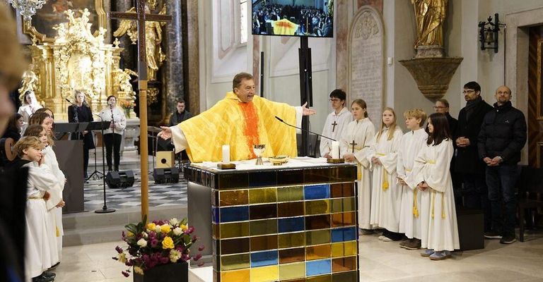 Ein Priester in einer gelben Robe leitet einen Gottesdienst mit einem Chor. Kerzen und Blumen schmücken den Altar. Menschen sehen auf einer großen Leinwand zu.