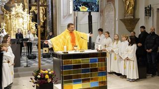 Ein Priester in einer gelben Robe leitet einen Gottesdienst mit einem Chor. Kerzen und Blumen schmücken den Altar. Menschen sehen auf einer großen Leinwand zu.
