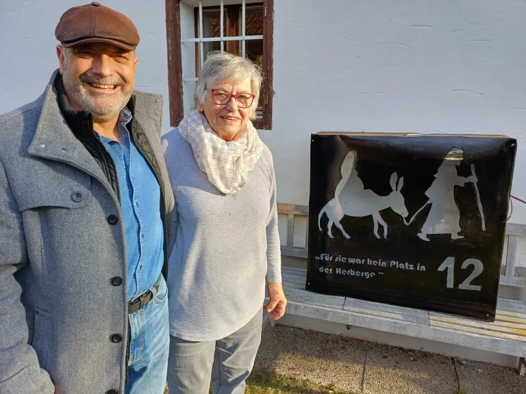 Ein Mann und eine Frau stehen vor einem weißen Gebäude mit Fenster und Bank. Sie lächeln. Die Frau trägt einen Schal. Hinter ihnen befindet sich ein schwarzes Schild mit einer Silhouette eines Esels und einer Person.