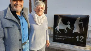 Ein Mann und eine Frau stehen vor einem weißen Gebäude mit Fenster und Bank. Sie lächeln. Die Frau trägt einen Schal. Hinter ihnen befindet sich ein schwarzes Schild mit einer Silhouette eines Esels und einer Person.