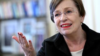 Eine ältere Frau mit kurzen Haaren, die eine Brille und eine schwarze Jacke trägt, lächelt und gestikuliert mit ihrer Hand, wahrscheinlich in einem Interview-Setting mit Bücherregalen im Hintergrund.