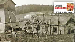 Ein Schwarz-Weiß-Bild eines Dorfes mit Häusern, einem Turm und einem Wald im Hintergrund. Rechts ist ein Schild mit dem Text 'Aggratt Burgenland' zu sehen.
