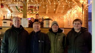 Vier Menschen posieren für ein Foto in einem warm beleuchteten Raum mit Holzwänden und Lichterketten. Sie lächeln und hinter ihnen stehen zwei Frauen.