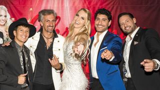 Vier Personen posieren für ein Foto vor einem rosa Hintergrund, lächeln und zeigen. Die Frau trägt ein schimmerndes Kleid.