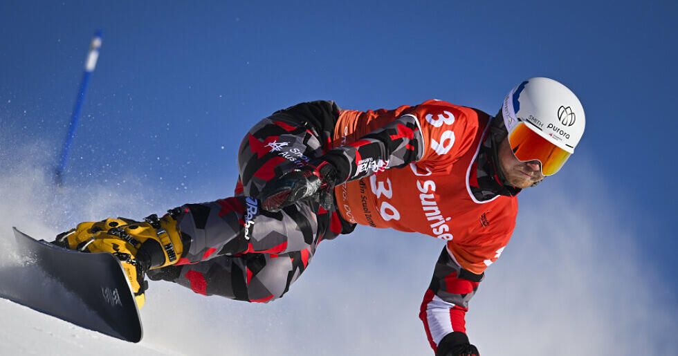 Ein Snowboarder führt auf einem verschneiten Berg einen Trick aus, trägt einen roten Anzug mit der Nummer 38 und einen weißen Helm.