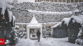 Eine Person in einem schwarzen Mantel geht durch einen verschneiten Bereich mit einem weißen Zelt, das mit Schnee bedeckt ist.