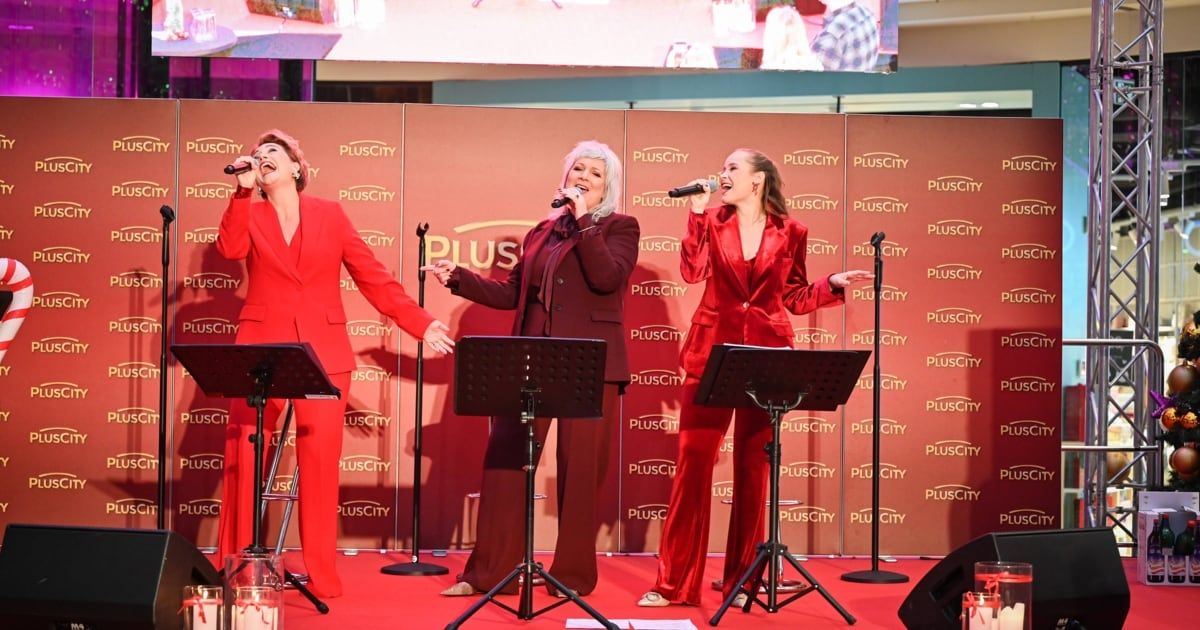 Drei Frauen in roten Anzügen stehen auf der Bühne und singen mit Mikrofonen. Sie haben eine Kulisse mit dem PlusCity-Logo im Hintergrund.