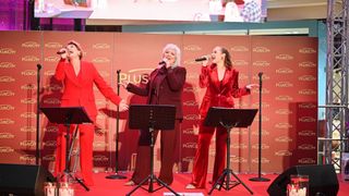 Drei Frauen in roten Anzügen stehen auf der Bühne und singen mit Mikrofonen. Sie haben eine Kulisse mit dem PlusCity-Logo im Hintergrund.