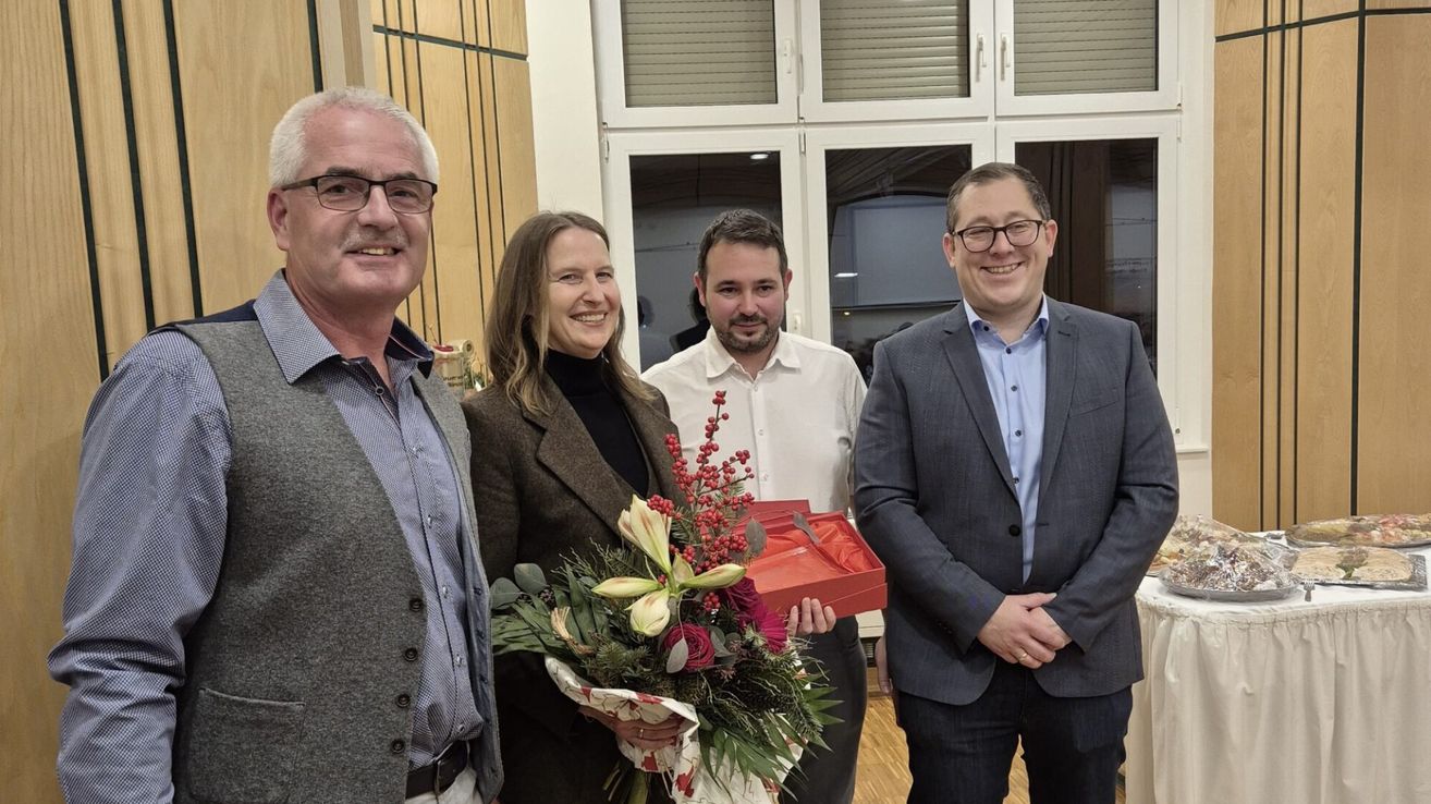 Vier Personen stehen zusammen in einem Raum, eine Frau hält einen Blumenstrauß und eine rote Box. Die Leute lächeln und scheinen für ein Foto zu posieren. Der Mann auf der linken Seite trägt eine Brille.