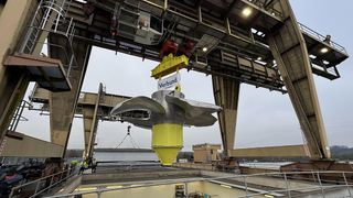 Ein großer gelber und grauer Propeller wird von einem Kran mit einem Schild 'Verbund' gehoben. Arbeiter stehen auf der Anlegestelle, und ein Gebäude mit einem Turm ist in der Ferne.