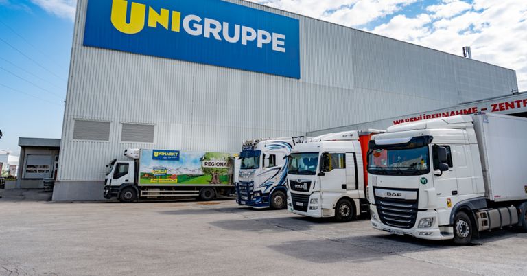 Mehrere LKWs sind vor einem UNIGRUPE-Lager geparkt. Die LKWs haben Werbung für Unimarkt und MAN. Der Himmel ist bewölkt.