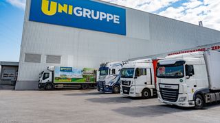 Mehrere LKWs sind vor einem UNIGRUPE-Lager geparkt. Die LKWs haben Werbung für Unimarkt und MAN. Der Himmel ist bewölkt.