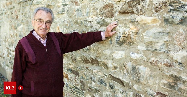 Ein älterer Mann mit Brille und einem bordeauxroten Pullover steht lächelnd vor einer Steinmauer.