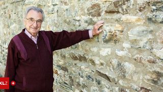 Ein älterer Mann mit Brille und einem bordeauxroten Pullover steht lächelnd vor einer Steinmauer.