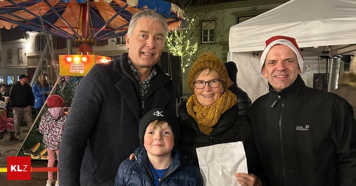 Vier Personen stehen vor einem Weihnachtsmarkt. Ein Junge hält ein Papier, und eine Frau trägt eine gestrickte Mütze.