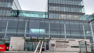 Das Bild zeigt den Eingang zum Justizzentrum Leoben, einem modernen Gebäude mit Glaswänden und einem auffälligen Schild. Der Eingang wird von Metallpfosten flankiert, und der Bereich ist sauber und gut gepflegt.